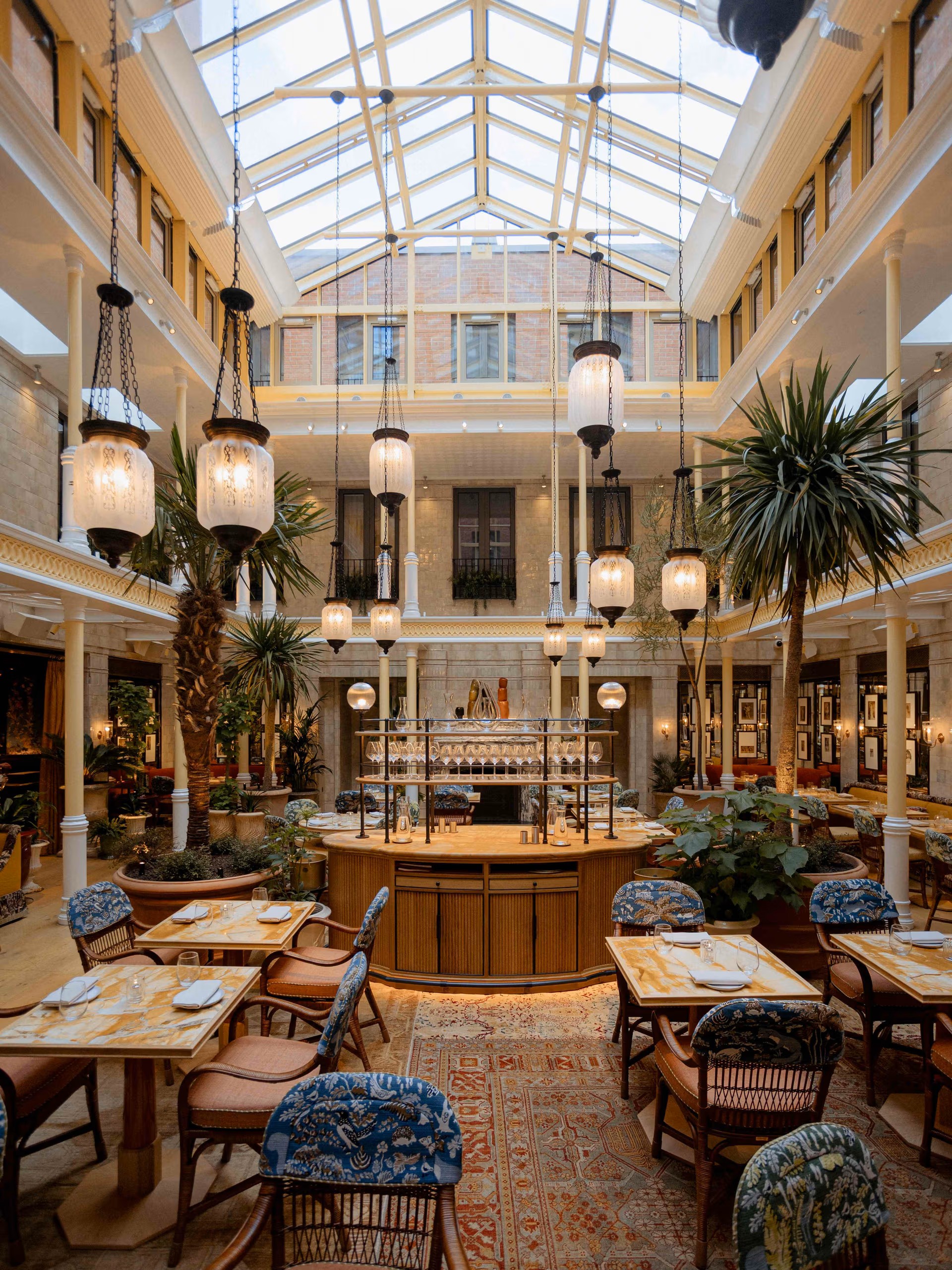 An hotel's atrium boasts ornately decorated interiors with live palm trees, wood and fabric furnishings, a central, glamorous bar, and pendant lanterns in wrought iron and glass.