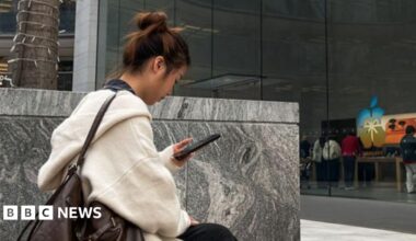Tim Cook with short white hair, walking, flashing the peace sign wearing a black blazer and white collared shirt.