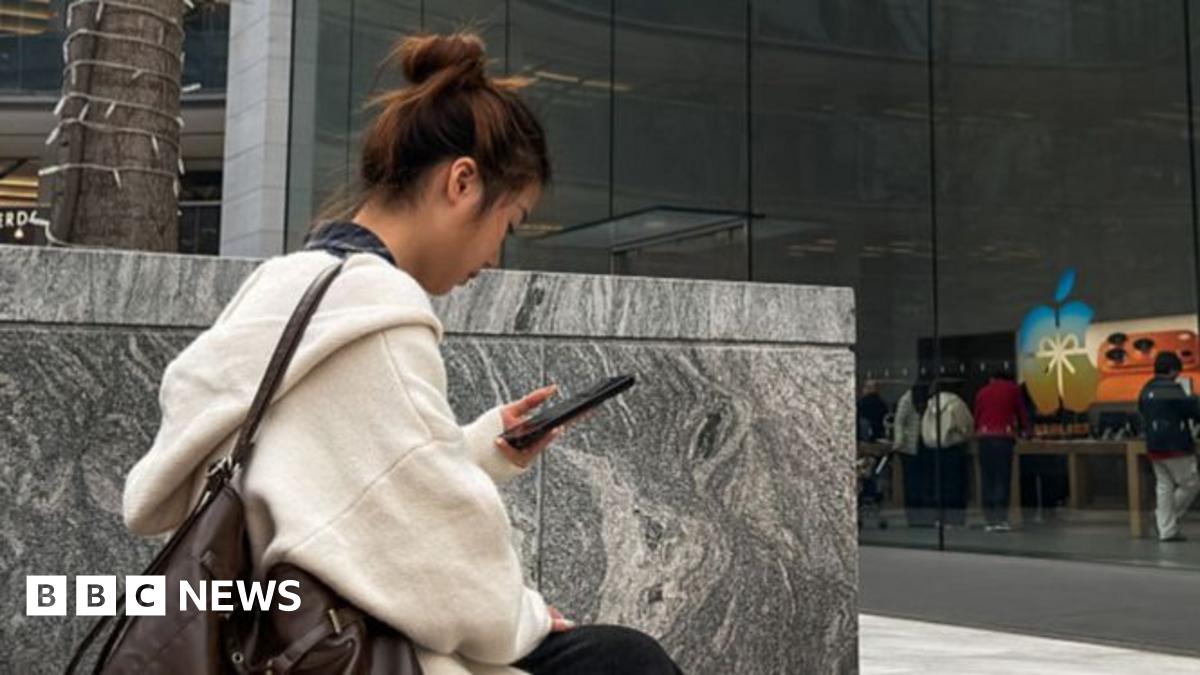 Tim Cook with short white hair, walking, flashing the peace sign wearing a black blazer and white collared shirt.