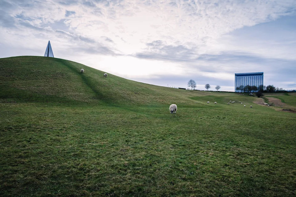 A serene winter landscape of Campbell Park in Milton Keynes, featuring a small monument on a curvy hill, sheep grazing on a green slope, and a multist