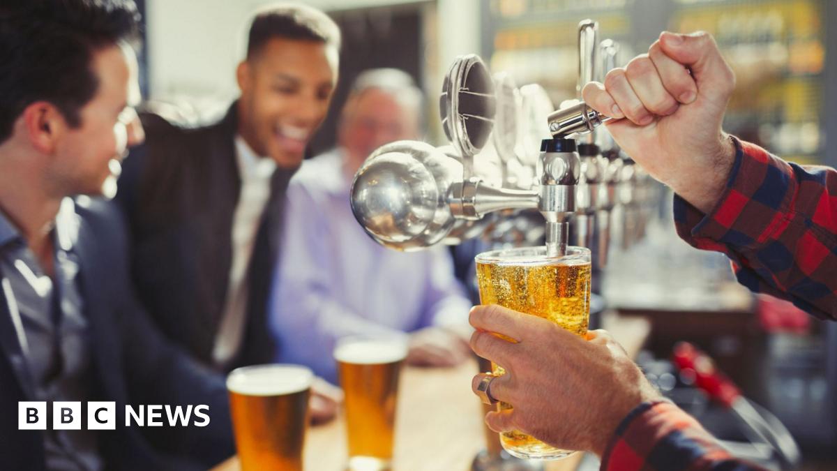 Close up of a pint being pulled from a tap. In the background there are blurred images of young men in shirts and jackets sitting at the bar with full pints in front of them. They look cheerful.