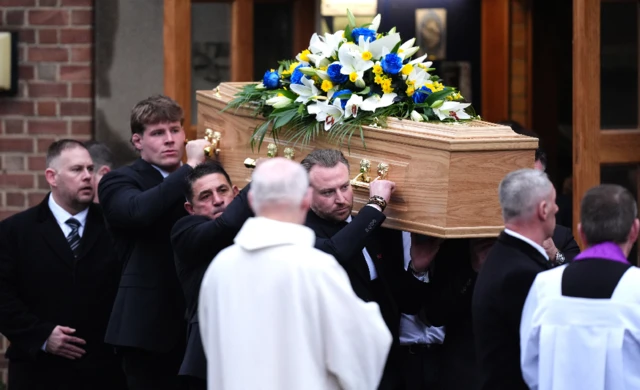 Image showing the bearers carrying Yorath's coffin out of the church. They are wearing black suits and the coffin has a floral tribute on top.