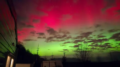 BBC Weather Watchers/GeorgePhoenix Green and red aurora light is visible above a tree line and scattered clouds, with the side of a building and several parked objects visible in the foreground.