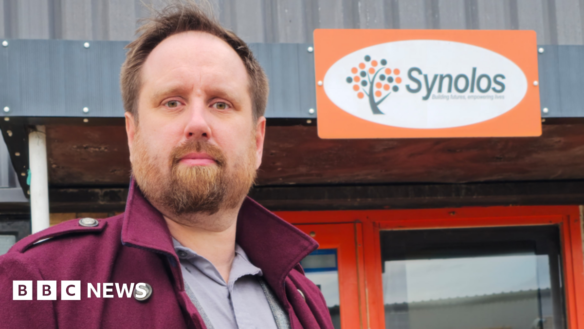 Barry Ingleton, a man with brown hair and a full beard, looks at the camera. He is wearing a maroon coat with an upturned collar over a grey shirt. Behind him is a sign on the front of a building reads "Synolos" next to a stylized symbol representing a tree.