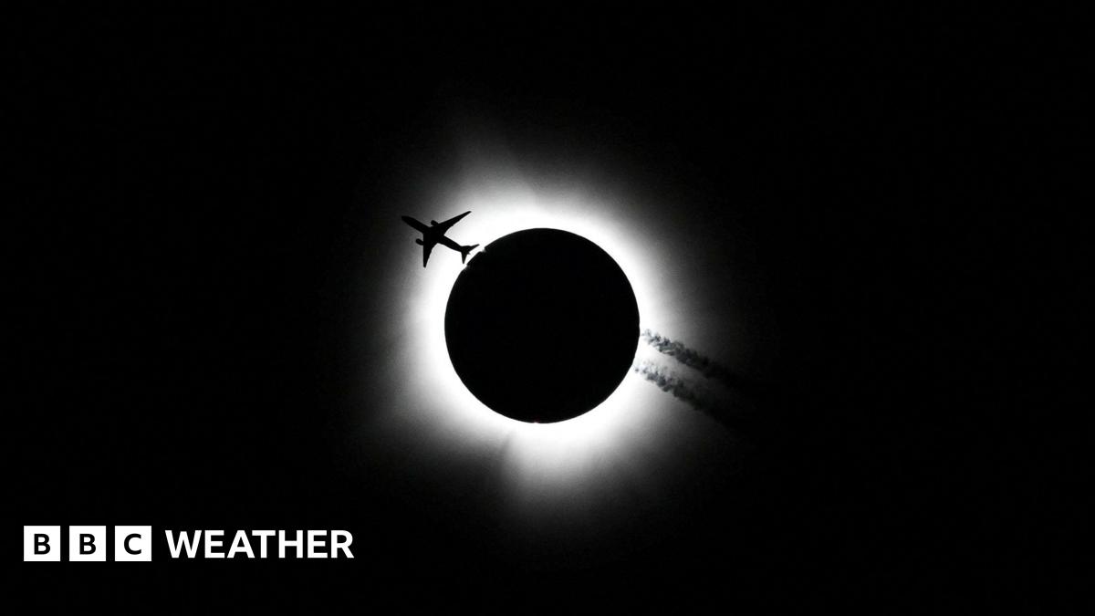 Black sky with white halo around a black disc. Silhouetted plane travelling through white halo