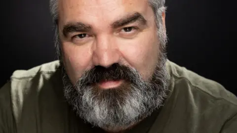 Ian Bustard A man with a black and grey beard wearing a khaki green shirt