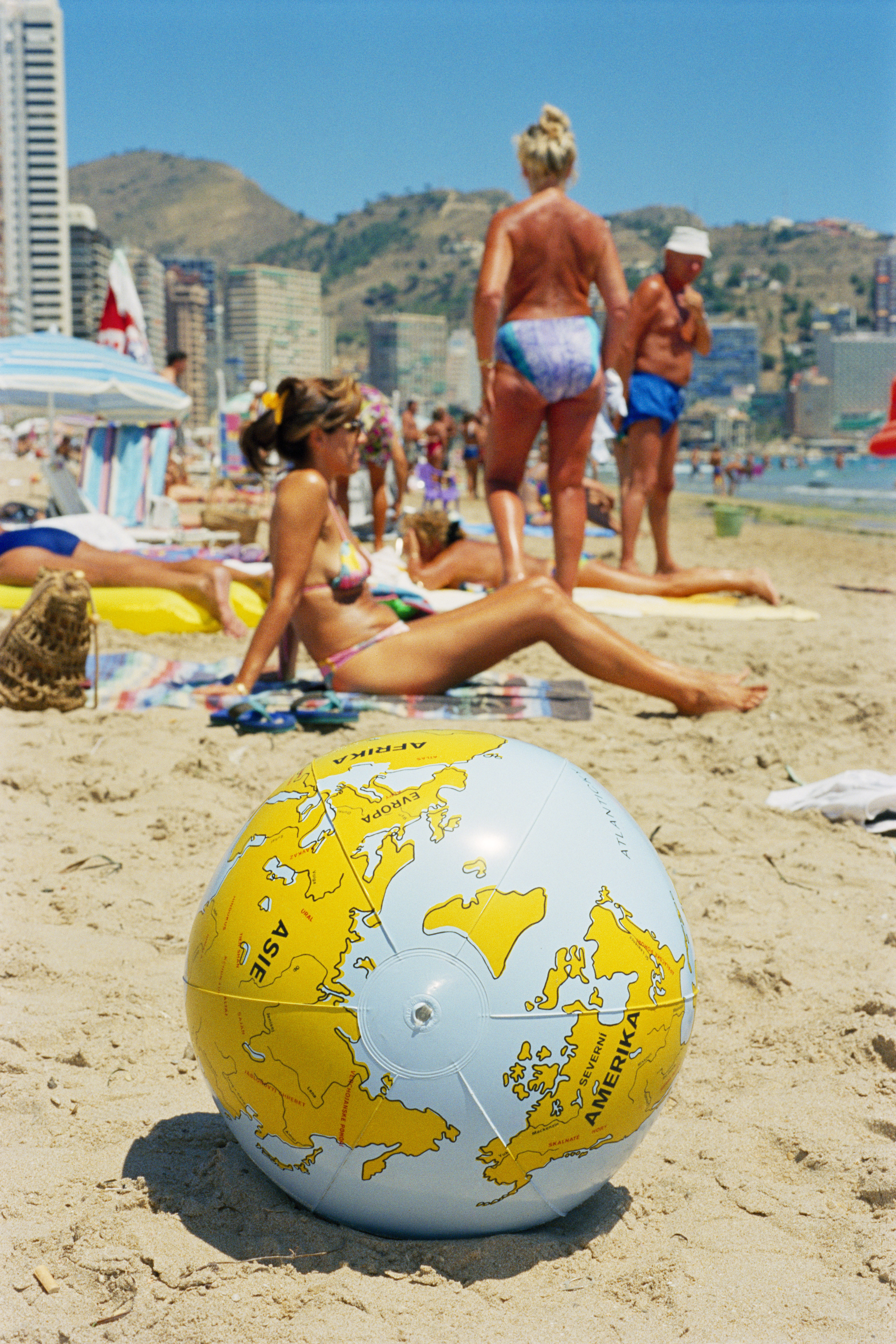 An inflatable beach ball designed like a world globe sits in the sand in the foreground, with sunbathers and a city skyline visible in the blurred background.