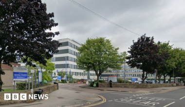 A black and white striped modern building in the background, with a row of trees and a road in the foreground. To the left is a blue and white sign which says "Barnsley Hospital".