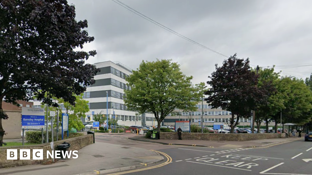 A black and white striped modern building in the background, with a row of trees and a road in the foreground. To the left is a blue and white sign which says "Barnsley Hospital".