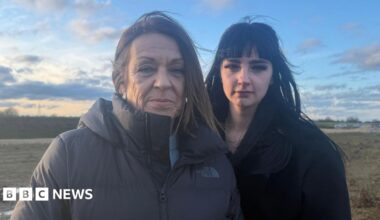 Jessica and Nicole Groucott are standing next to each other, Nicole on the left is wearing a black zipped up coat and she has medium length brown hair. Jessica, who is next to her, has long black hair and is wearing a black coat. The pair are in a field and the skies behind them are filled with clouds while sunset descends.