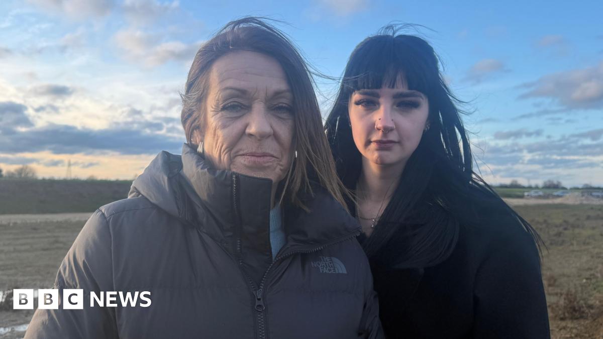 Jessica and Nicole Groucott are standing next to each other, Nicole on the left is wearing a black zipped up coat and she has medium length brown hair. Jessica, who is next to her, has long black hair and is wearing a black coat. The pair are in a field and the skies behind them are filled with clouds while sunset descends.