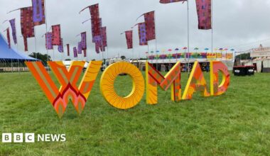 Womad sign at the festival grounds. The abbreviation is spelled out with large, orange and yellow printed letters on grass. The festival site can be seen behind in the distance - there are colourful flags, tents and stands.