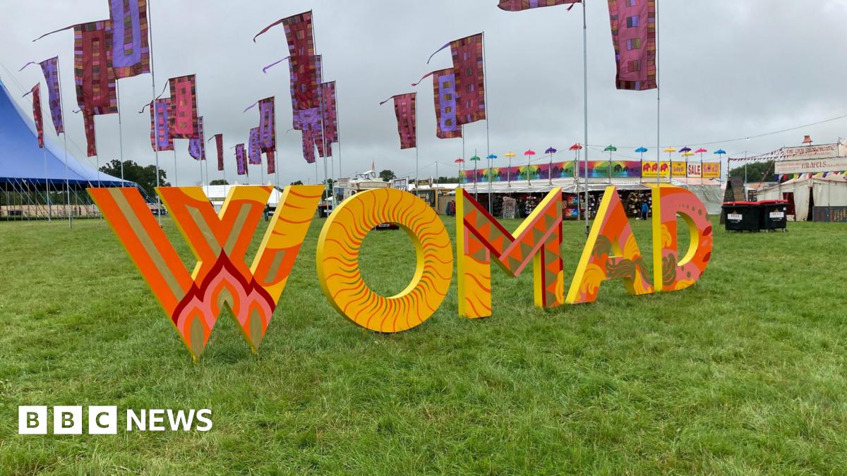 Womad sign at the festival grounds. The abbreviation is spelled out with large, orange and yellow printed letters on grass. The festival site can be seen behind in the distance - there are colourful flags, tents and stands.