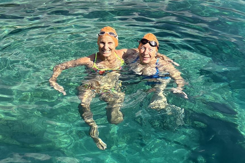 Two woman in bathing suits and swimming caps float in clear blue water with boats and a rocky shore in the background.