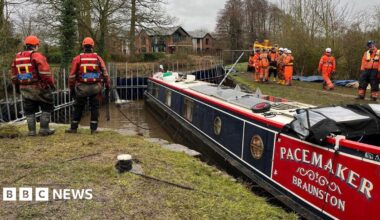 dark blue and red canal boat in a small section of canal blocked off with metal dams. it is in water and there are dozens of people standing on banks either side of it in orange outfits