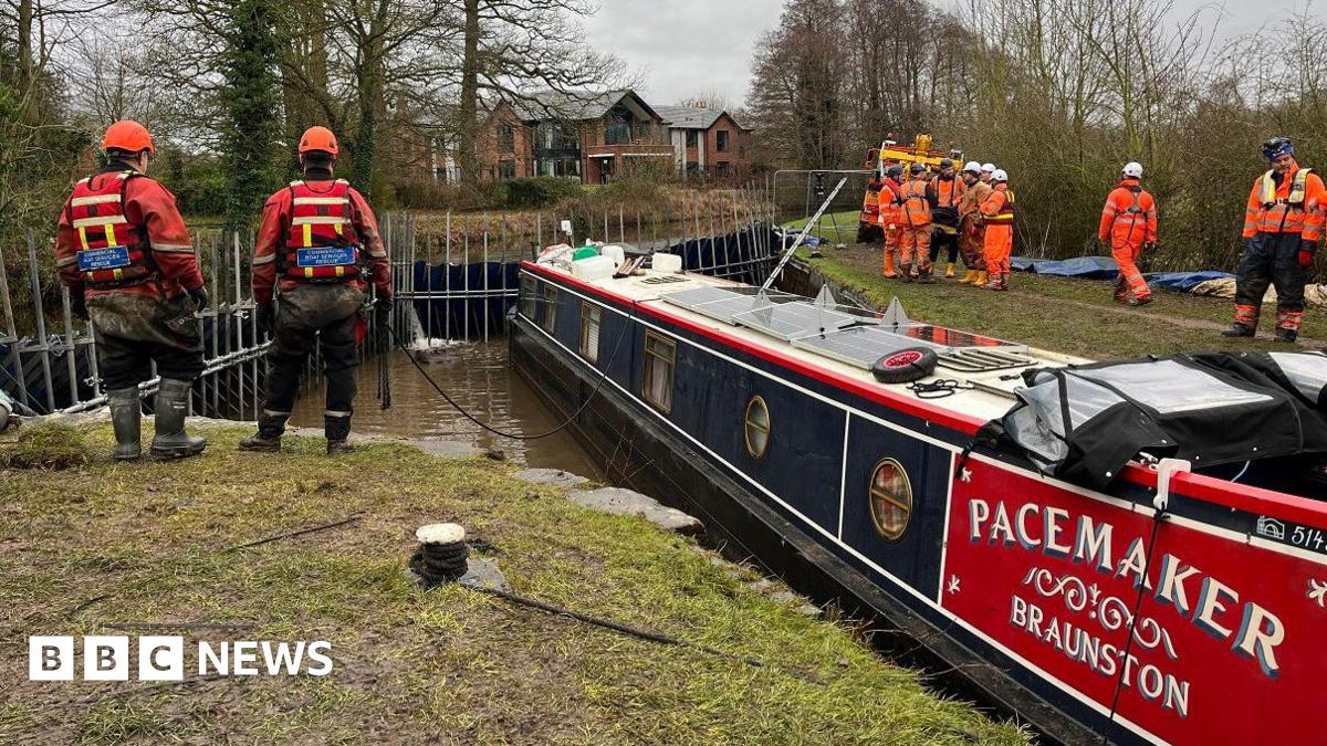 dark blue and red canal boat in a small section of canal blocked off with metal dams. it is in water and there are dozens of people standing on banks either side of it in orange outfits
