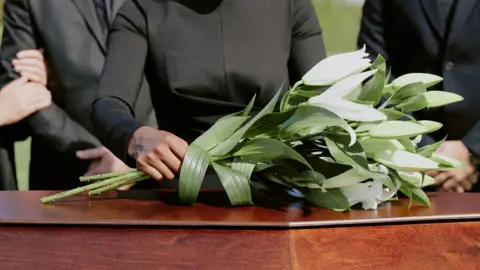 Getty Images Stock photo shows white flowers being placed on a wooden coffin by people attending a funeral outdoors.