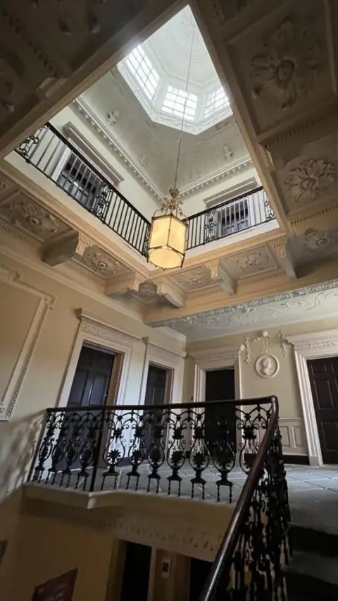Susan Amaku, Woodhall Planning and Conservation The stairway and upper hall of a Georgian townhouse, with decorative plasterwork on walls and ceilings, and an octagonal lantern in the roof. The walls are cream-coloured and there are four black doors. The staircase has a black, iron balustrade with an intricate design.