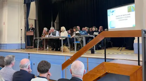 A man at a plinth is talking while many others sit next to him on a stage