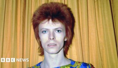An image of David Bowie from the 1970s, he is standing against a yellow curtain and has dark red hair.