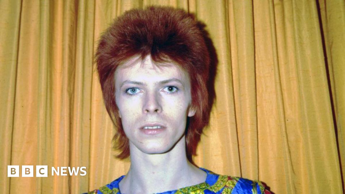 An image of David Bowie from the 1970s, he is standing against a yellow curtain and has dark red hair.