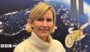 This is a head and shoulders picture of Kata Escott. She is in the centre of the frame, smiling and looking at the camera. Behind her is a satellite image of the earth from space at night, with lights visible on the land masses. An image of a satellite can also be seen on the wall behind her.
