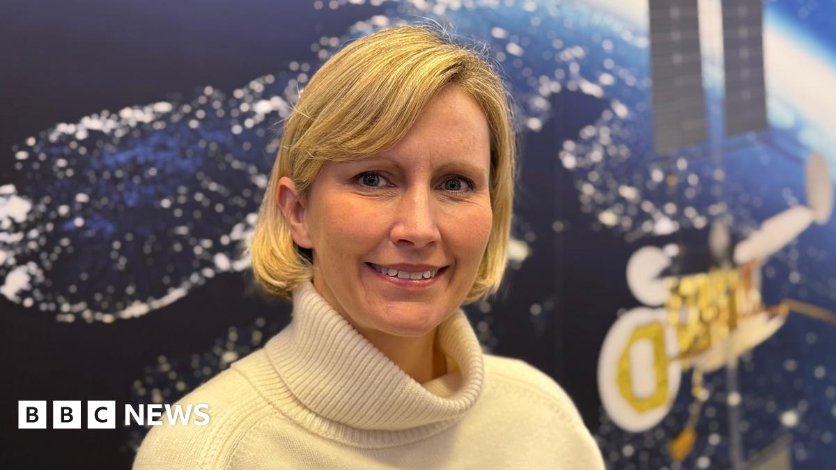 This is a head and shoulders picture of Kata Escott. She is in the centre of the frame, smiling and looking at the camera. Behind her is a satellite image of the earth from space at night, with lights visible on the land masses. An image of a satellite can also be seen on the wall behind her.