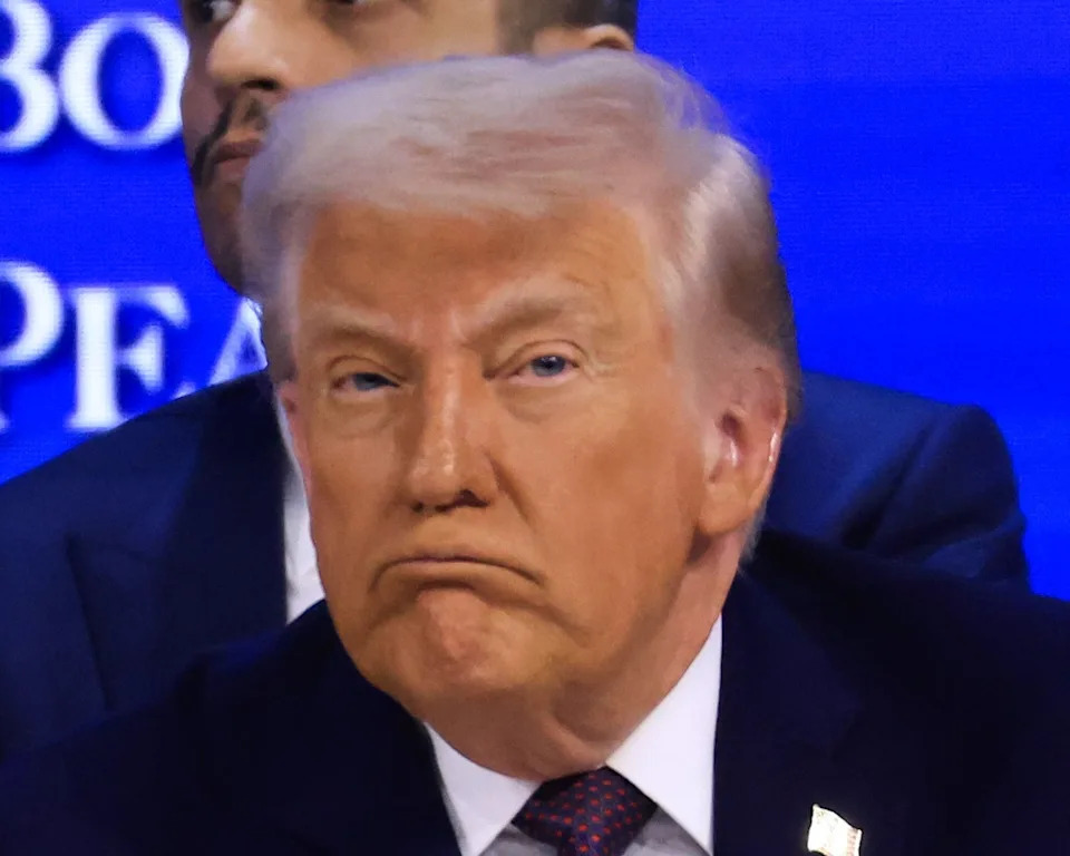 A man in a suit whispers to another suited man seated next to him at a formal event, with others in business attire in the background
