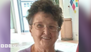 Celia Adams is pictured smiling at the camera wearing glasses and a purple top. She has short brown hair.