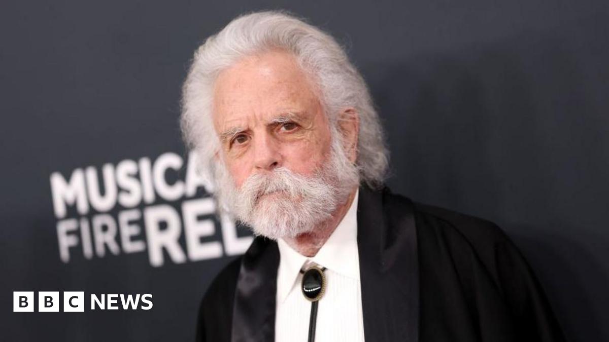 Bob Weir, with a dark suit and shite shirt, has a white beard and moustache