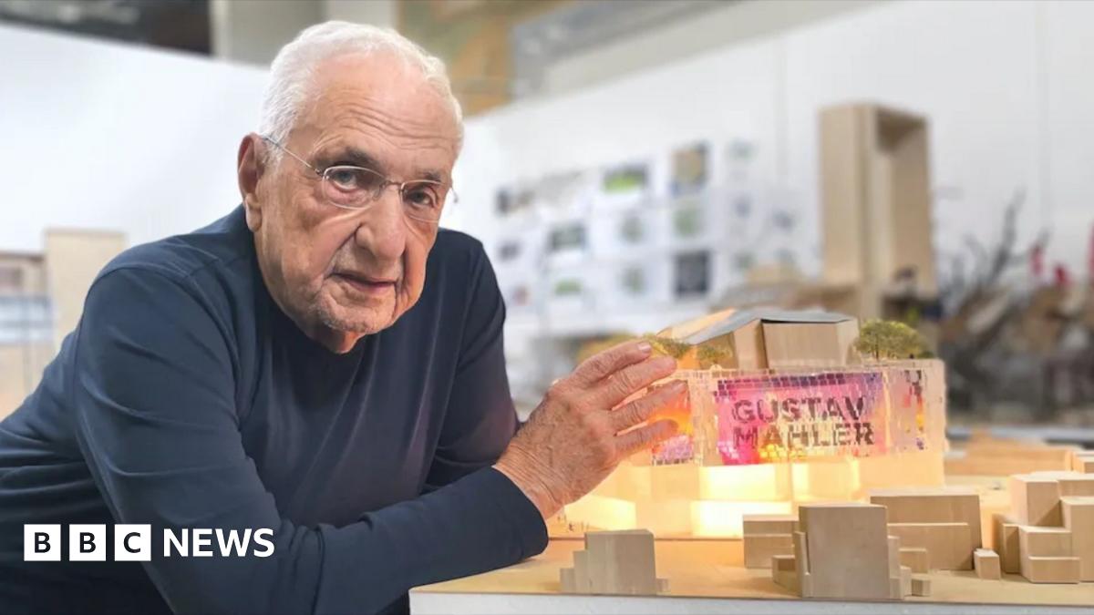 A person wearing a dark long-sleeve shirt is leaning on a table, presenting a detailed architectural model, featuring illuminated sections, miniature trees, and geometric structures in a modern studio setting.