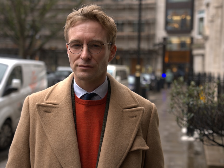 A man wearing thin-framed glasses and a tan jacket looks seriously at the camera while standing on a wet footpath in a city