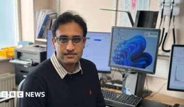 A doctor wearing a blue jumper and glasses is sat in his consulting room staring straight into the camera. He is surrounded by computer monitors and a brown wooden desk. A stack of paperwork is visible in the background.