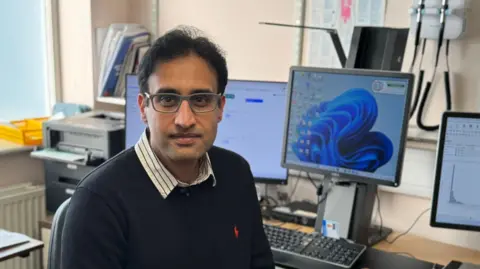 BBC A doctor wearing a blue jumper and glasses is sat in his consulting room staring straight into the camera. He is surrounded by computer monitors and a brown wooden desk. A stack of paperwork is visible in the background.
