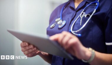 A stock image of a nurse in a navy blue uniform on a tablet with a stethoscope around their neck. Their face is not in view.