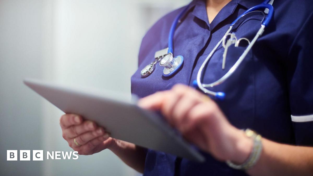 A stock image of a nurse in a navy blue uniform on a tablet with a stethoscope around their neck. Their face is not in view.