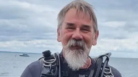 The Seahorse Trust Nick Garrick-Maidment smiling at the camera and wearing diving gear. He has grey beard and hair. He is stood in front of the sea. 
