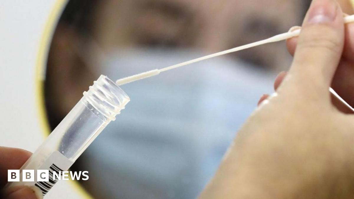 A close-up of the hands of a person placing a cotton swab into a clear test tube in front of a small mirror. In the reflection, you can see the blurred face of the same person wearing a blue mask.