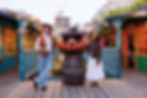 Cowboy and woman smiling, standing by a large pumpkin decoration with hats, holding drinks. Festive lights and colorful buildings in the background.