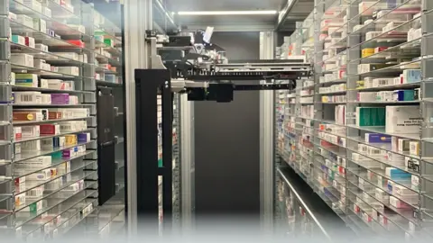 Matt Knight/BBC A black robotic device is positioned in the middle of a room. On either side are shelves stacked with various types of medication. There is a dark grey door at the far end of the room behind the robot.