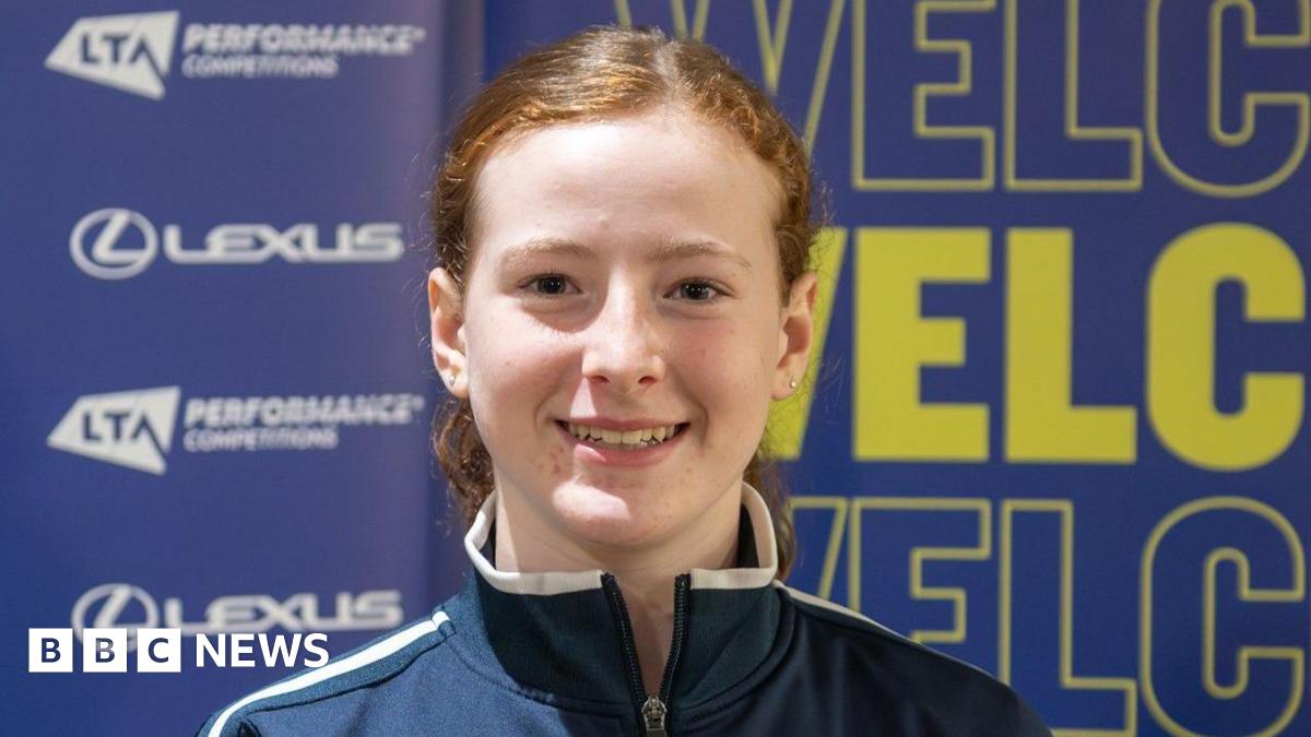 A girl smiles at the camera in a head and shoulders image. She has brown eyes and auburn hair tied back and is wearing a navy and white zip-up sports top. The background is blue and a sponsors' wall covered in yellow and blue wording, including Lexus and LTA Performance Competition.