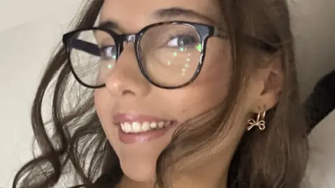 PA Media A close-up picture of Amber looking at the camera. She has long dark curled hair with blue eyes and dark-rimmed glasses.