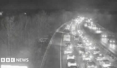 A traffic camera image above the M11 motorway. It is night time and the vehicles on both sides of the carriageway have their lights on. The carriageway closest to the camera has long queues of traffic.
