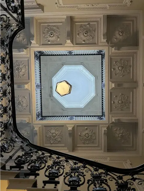 Susan Amaku, Woodhall Planning and Conservation A view looking straight up from a staircase towards a ceiling and lantern roof light. The staircase has an intricate black, iron balustrade. The ceiling has cream-coloured plasterwork, with rose designs. The lantern is eight-sided and appears light blue. There is a yellow ceiling light.