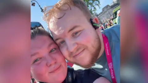 Richard Gallimore A close up selfie of Richard with his girlfriend, Sophie, at the Race for Life running event. Sophie, on the left, has brown hair and wears sunglasses on the top of her head. Richard is standing next to Sophie with his cheek touching her cheek. He wears a green t-shirt, a pink medal, earphones and has short blonde hair. 