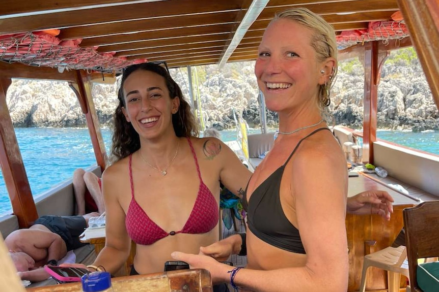 Ilgen Ilter and Aoife Doran wear bikinis and smile while on a boat near the coastline.