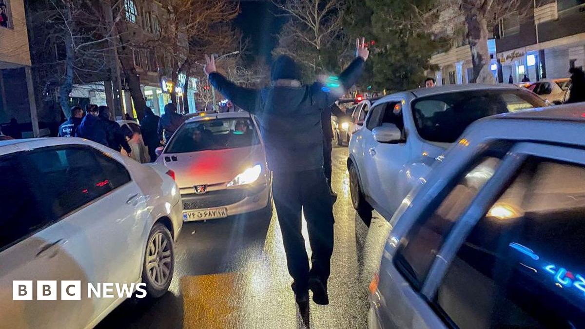 A person walks between moving cars on a road, while making the victory sign with his hands, at night-time in Hamedan, Iran (01/01/26)