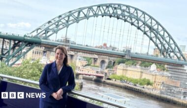 Rachel Reeves, who has dark brown hair, is wearing a navy blue suit and is standing on a balcony, leaning against a railing. She is smiling and facing the camera. Behind her is the green Tyne Bridge. It is almost fully visible with part of both the High Level Bridge and Swing Bridge in view behind it. Reeves stands on the Gateshead side of the river and part of Newcastle Quayside can also be seen.