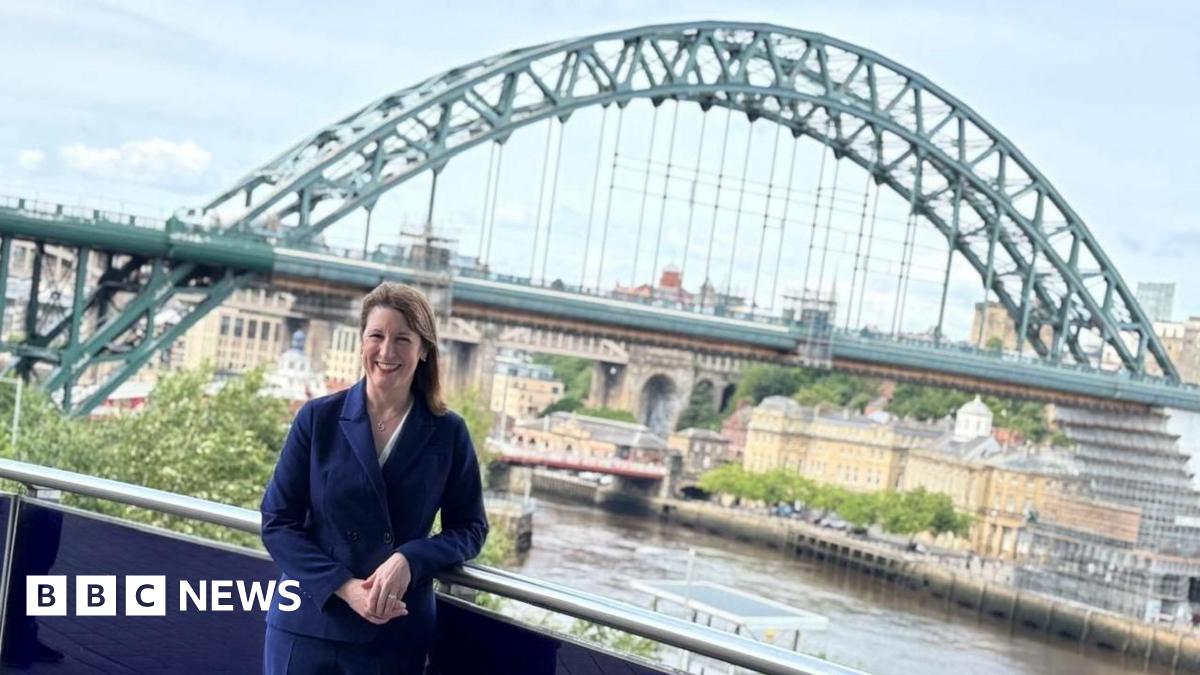 Rachel Reeves, who has dark brown hair, is wearing a navy blue suit and is standing on a balcony, leaning against a railing. She is smiling and facing the camera. Behind her is the green Tyne Bridge. It is almost fully visible with part of both the High Level Bridge and Swing Bridge in view behind it. Reeves stands on the Gateshead side of the river and part of Newcastle Quayside can also be seen.