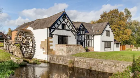 Matt Elson A ground-level outdoor view of the historic white watermill, featuring ornate black timber detailing and a steep slate roof. A large wooden waterwheel stands beside the flowing river, which runs past a stone retaining wall. The building is surrounded by neatly cut grass and mature autumn trees under a clear blue sky with scattered white clouds.